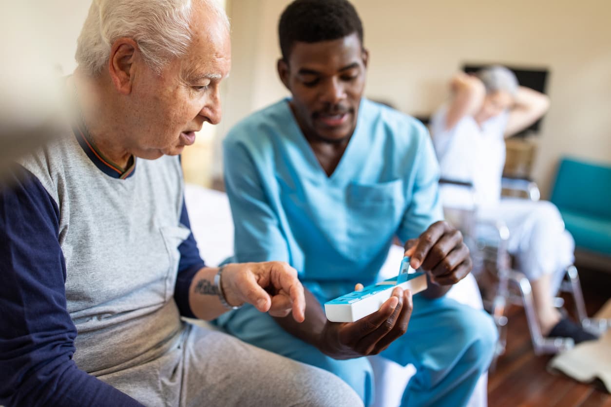 Male nurse sitting on bed and giving instructions to senior patient when to take his pills in retirement home.