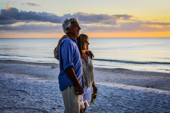 older adult husband and wife watching sunset on the beach with a bottle of wine